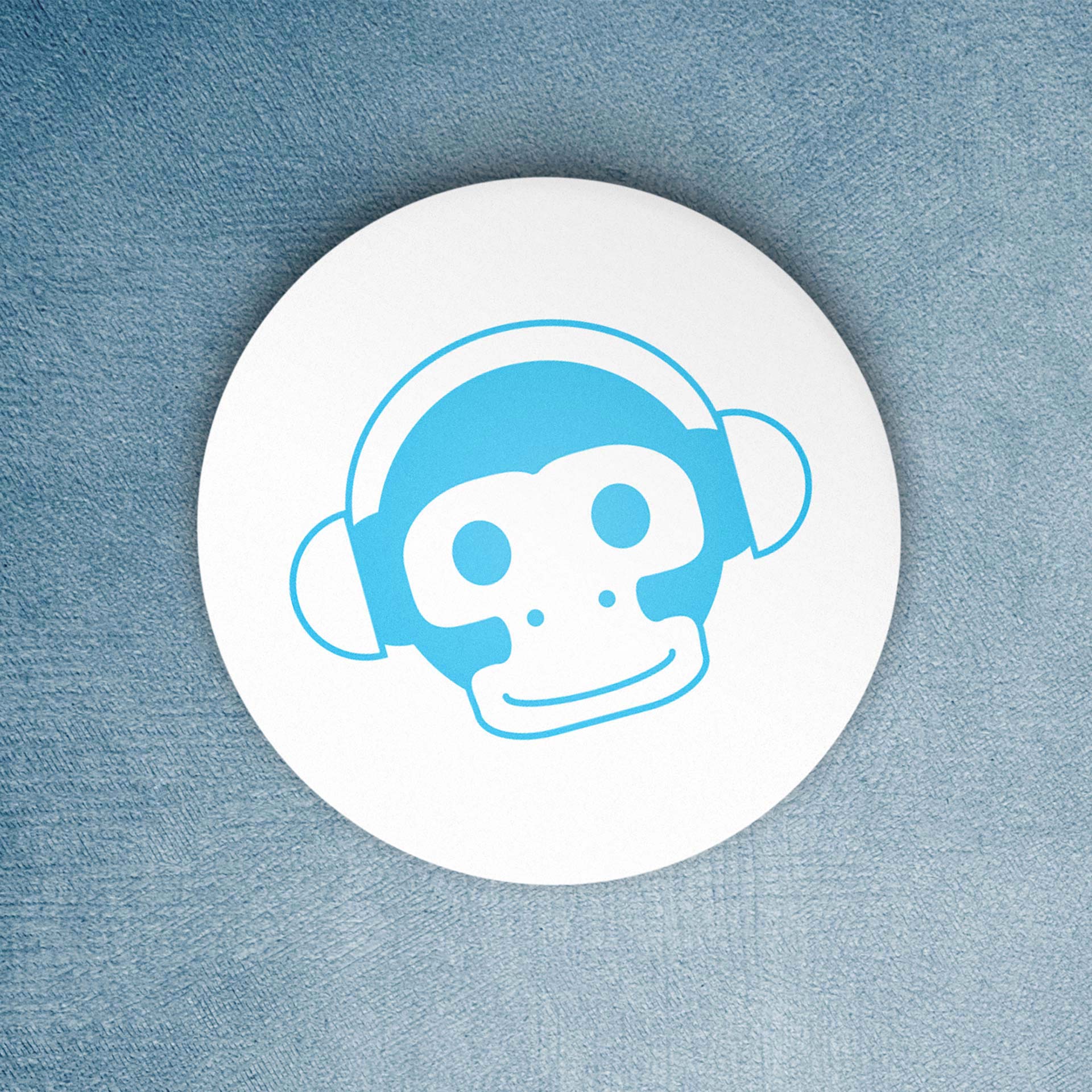 Sticker rond blanc posé sur une surface bleue, représentant une illustration stylisée en bleu d’un singe portant un casque audio.